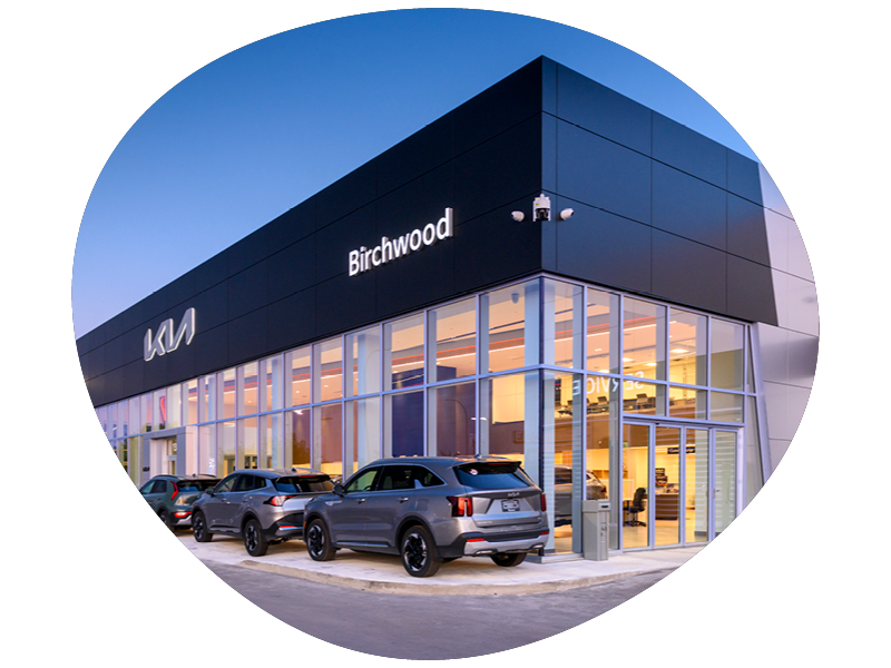 Modern Kia Birchwood car dealership building at dusk with several cars parked outside and large glass windows showcasing the interior.